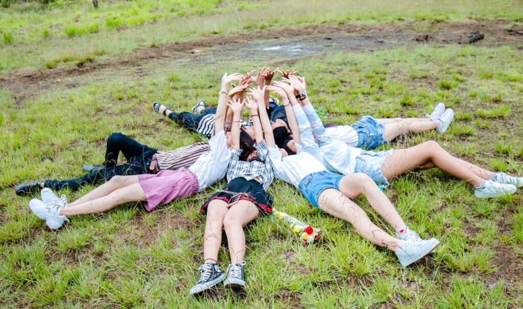 Young adults lying in a circle on grass, arms raised in a sign of joy and friendship outdoors.