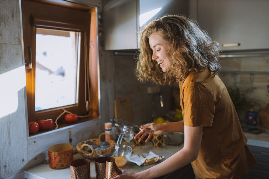 A happy woman with curly hair preparing food in a sunlit kitchen.