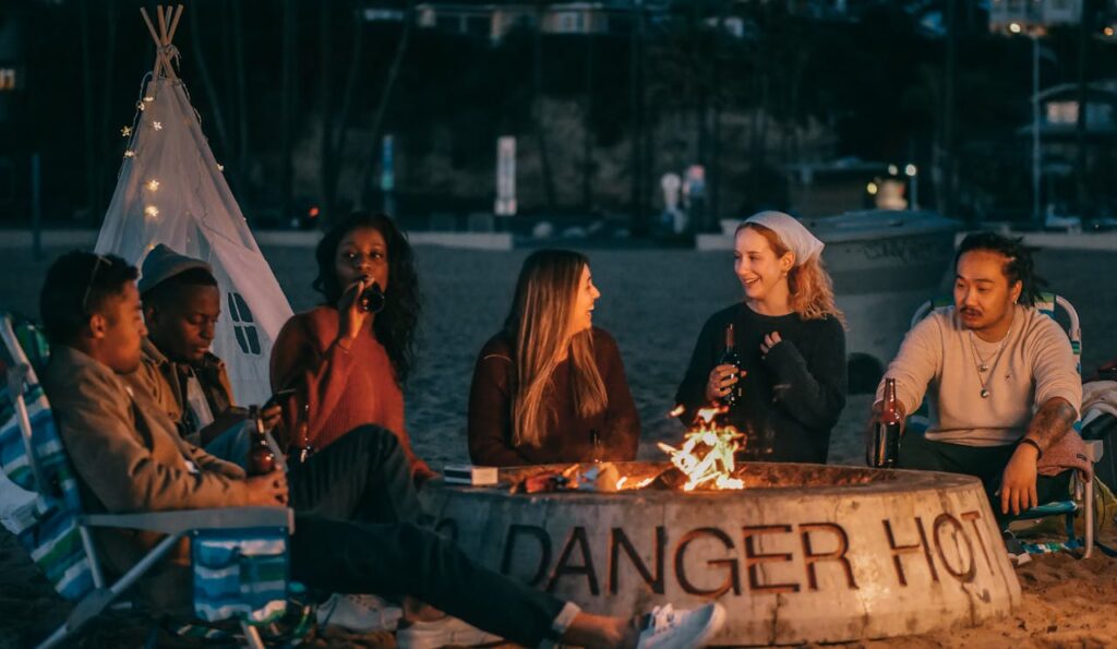 A diverse group of friends enjoying drinks and laughter around a beach bonfire at sunset.