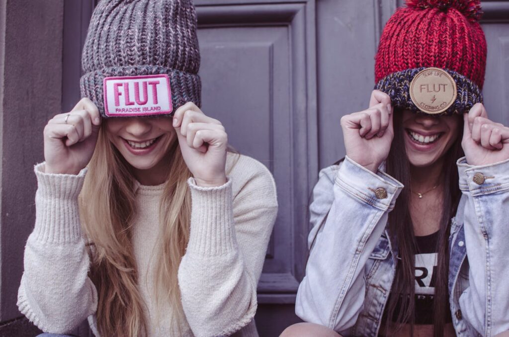 Two young women laughing and posing with colorful beanies, expressing joy and friendship.